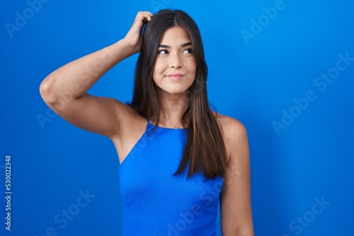 Hispanic woman standing over blue background confuse and wondering about question. uncertain with doubt, thinking with hand on head. pensive concept.