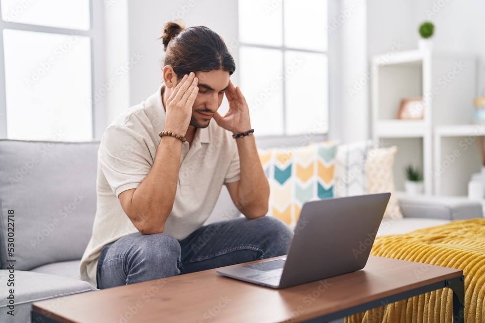 Young hispanic man stressed using laptop sitting on sofa at home