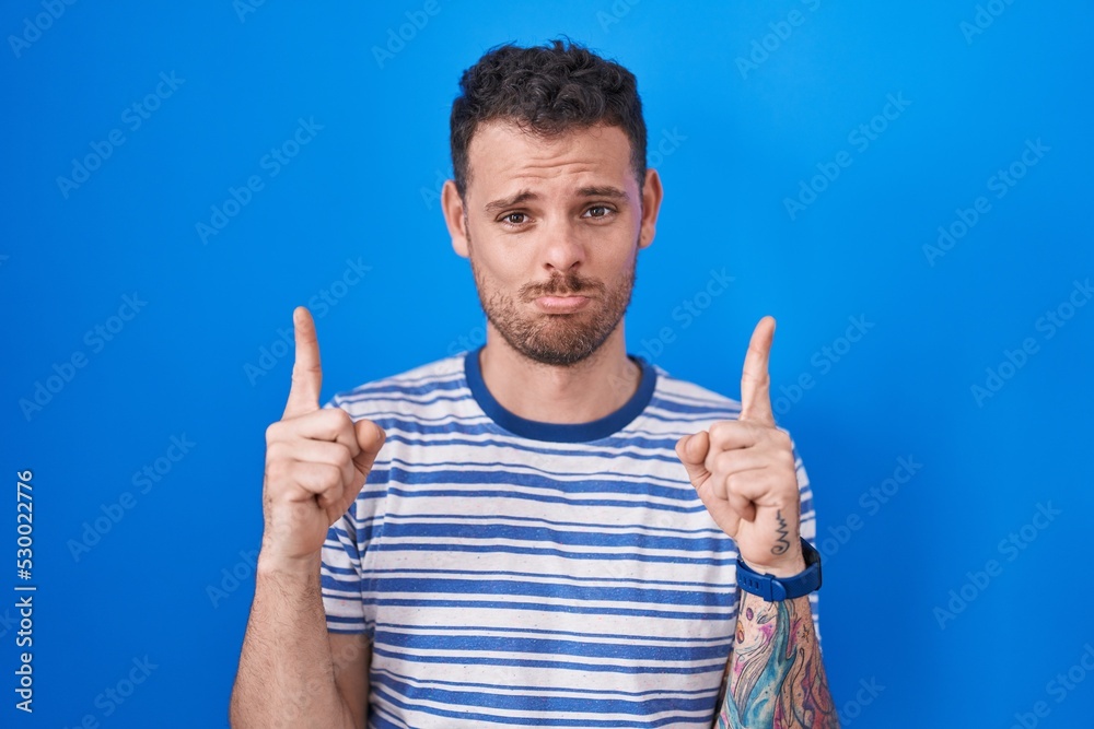 Young hispanic man standing over blue background pointing up looking sad and upset, indicating direction with fingers, unhappy and depressed. Young hispanic man standing over blue background pointing up looking sad and upset, indicating direction with fingers, unhappy and depressed.