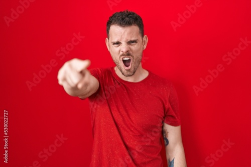 Young hispanic man standing over red background pointing displeased and frustrated to the camera, angry and furious with you