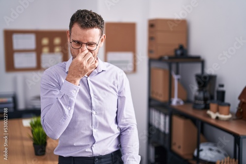 Young hispanic man at the office smelling something stinky and disgusting, intolerable smell, holding breath with fingers on nose. bad smell