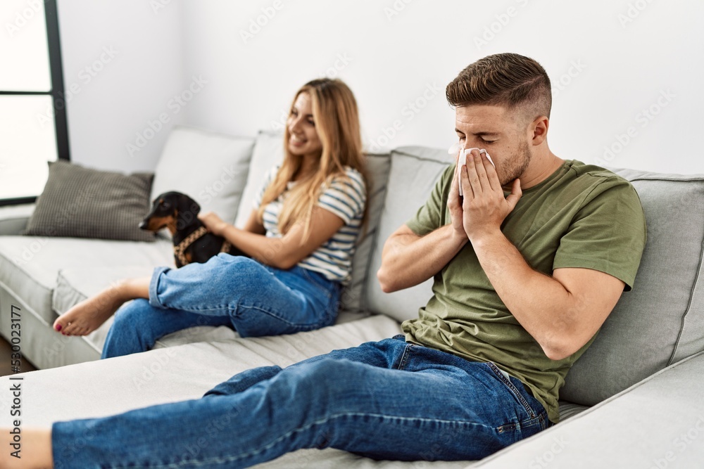 Young hispanic couple sitting on the sofa with dog at home. Man using handkerchief. Young hispanic couple sitting on the sofa with dog at home. Man using handkerchief.