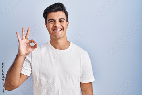 Hispanic man standing over blue background smiling positive doing ok sign with hand and fingers. successful expression. © Krakenimages.com