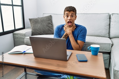 Young handsome hispanic man using laptop sitting on the floor shouting and suffocate because painful strangle. health problem. asphyxiate and suicide concept.