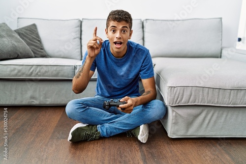 Young handsome hispanic man playing video game sitting by the sofa surprised with an idea or question pointing finger with happy face, number one