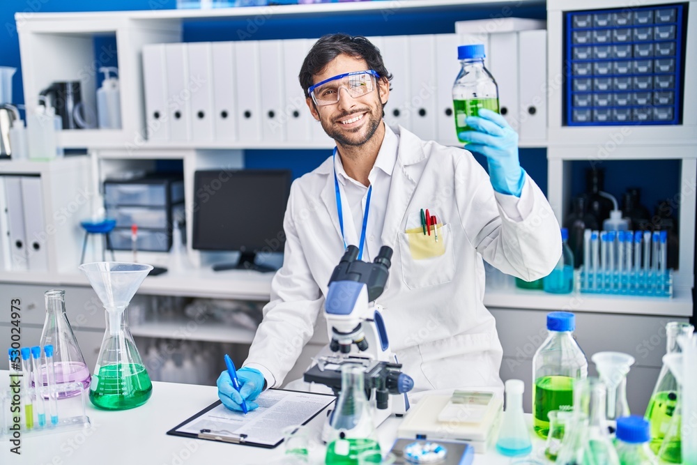 Young hispanic man scientist write on clipboard holding bottle at laboratory Young hispanic man scientist write on clipboard holding bottle at laboratory