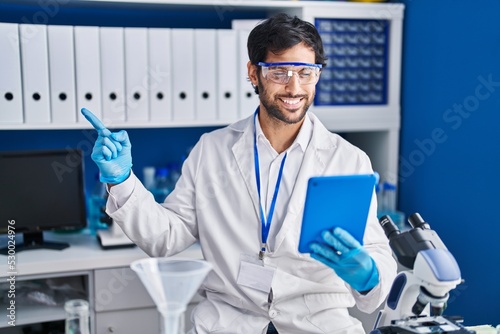 Handsome latin man working at scientist laboratory using tablet smiling happy pointing with hand and finger to the side