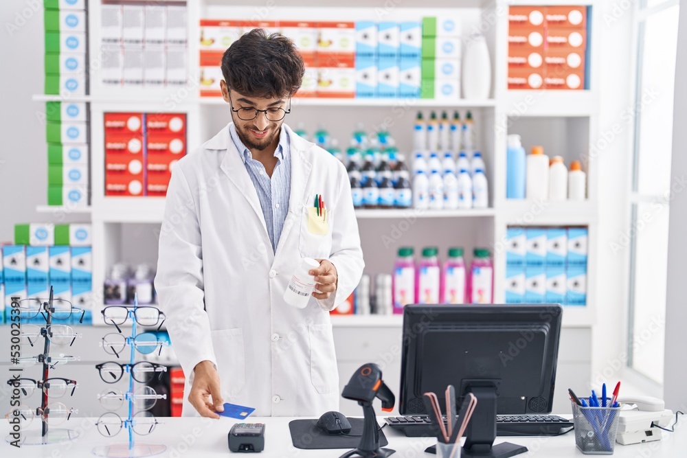 Young arab man pharmacist holding pills bottle using credit card at pharmacy Young arab man pharmacist holding pills bottle using credit card at pharmacy