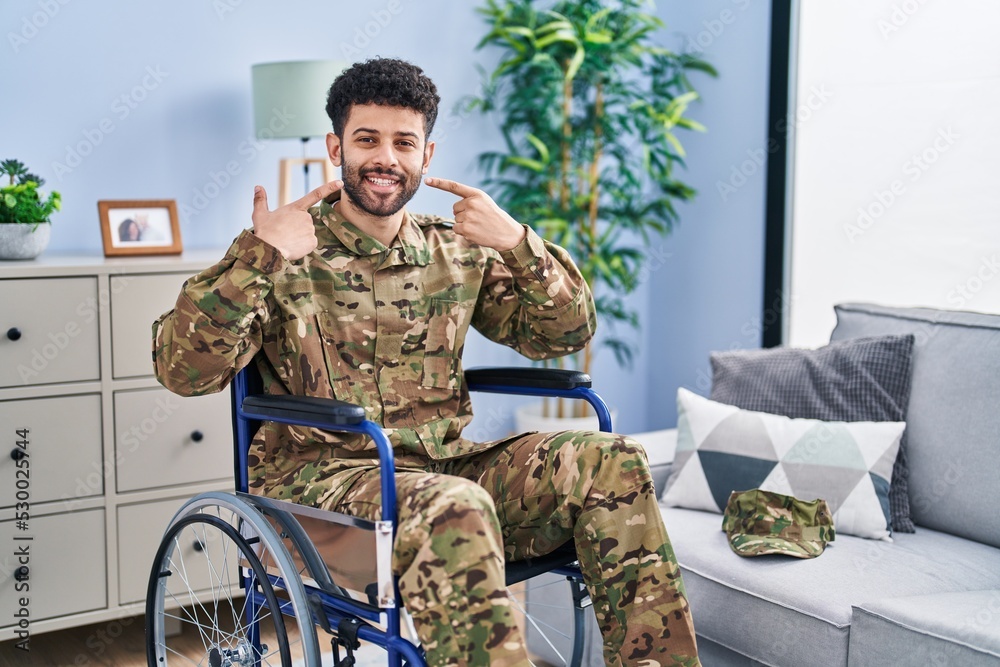 Fototapeta premium Arab man wearing camouflage army uniform sitting on wheelchair smiling cheerful showing and pointing with fingers teeth and mouth. dental health concept.
