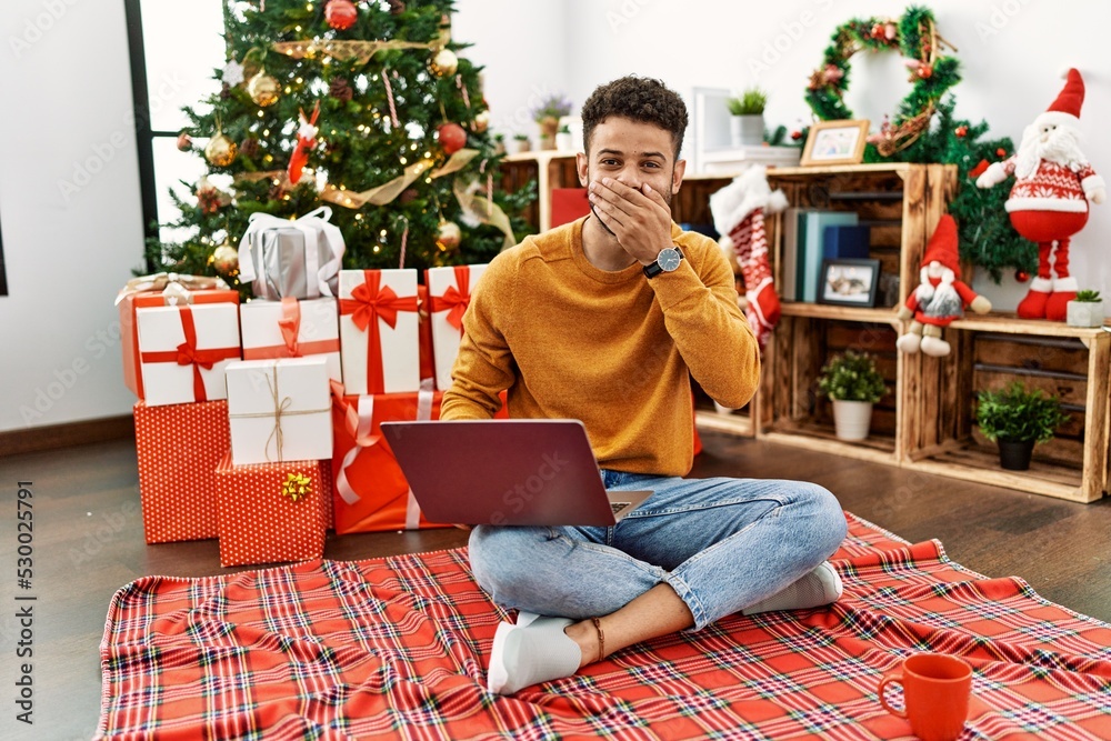Arab young man using laptop sitting by christmas tree laughing and embarrassed giggle covering mouth with hands, gossip and scandal concept Arab young man using laptop sitting by christmas tree laughing and embarrassed giggle covering mouth with hands, gossip and scandal concept
