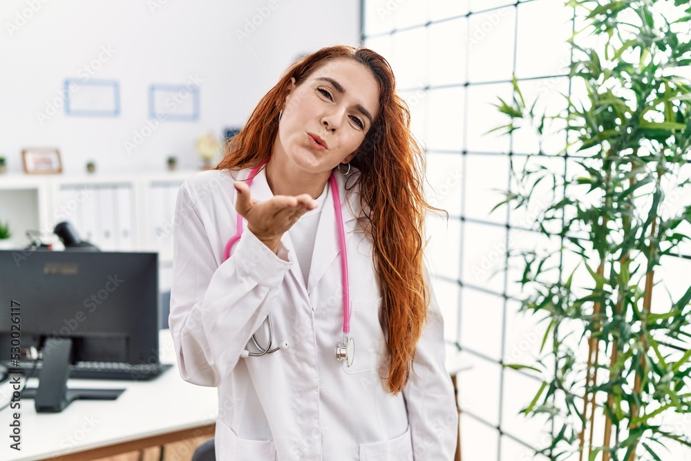 Young redhead woman wearing doctor uniform and stethoscope at the clinic looking at the camera blowing a kiss with hand on air being lovely and sexy. love expression. Young redhead woman wearing doctor uniform and stethoscope at the clinic looking at the camera blowing a kiss with hand on air being lovely and sexy. love expression.