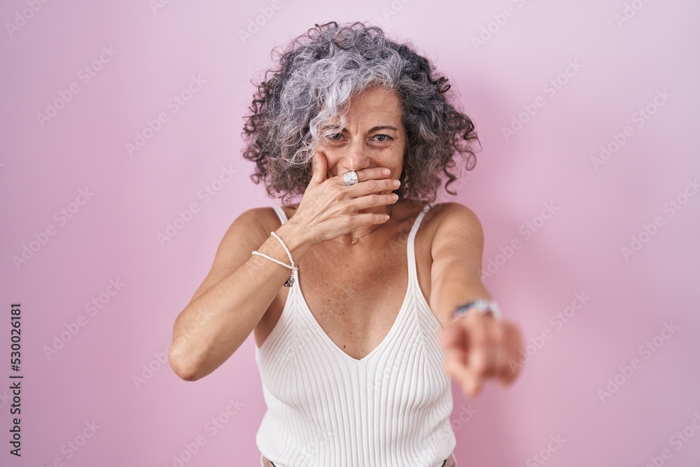 Middle age woman with grey hair standing over pink background laughing at you, pointing finger to the camera with hand over mouth, shame expression
