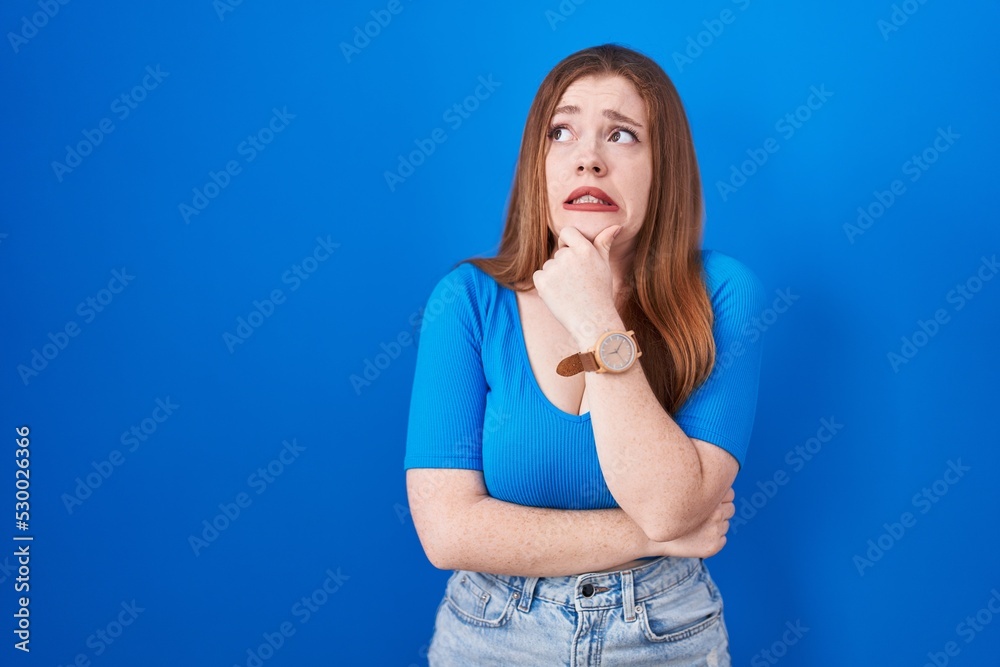 Redhead woman standing over blue background thinking worried about a question, concerned and nervous with hand on chin Redhead woman standing over blue background thinking worried about a question, concerned and nervous with hand on chin