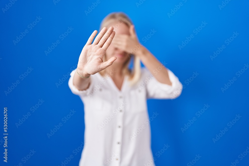 Young caucasian woman standing over blue background covering eyes with hands and doing stop gesture with sad and fear expression. embarrassed and negative concept. Young caucasian woman standing over blue background covering eyes with hands and doing stop gesture with sad and fear expression. embarrassed and negative concept.