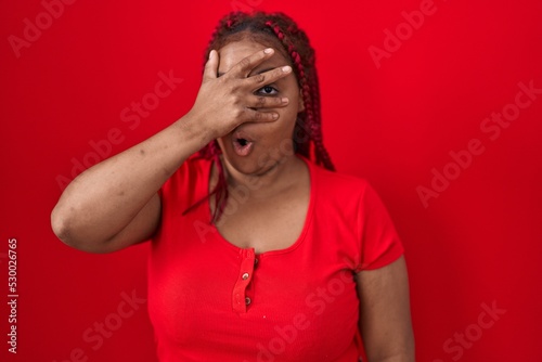 African american woman with braided hair standing over red background peeking in shock covering face and eyes with hand, looking through fingers with embarrassed expression.