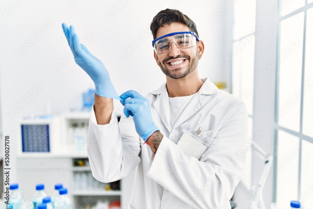 Young hispanic man working at scientist laboratory putting gloves on smiling with a happy and cool smile on face. showing teeth. Young hispanic man working at scientist laboratory putting gloves on smiling with a happy and cool smile on face. showing teeth.
