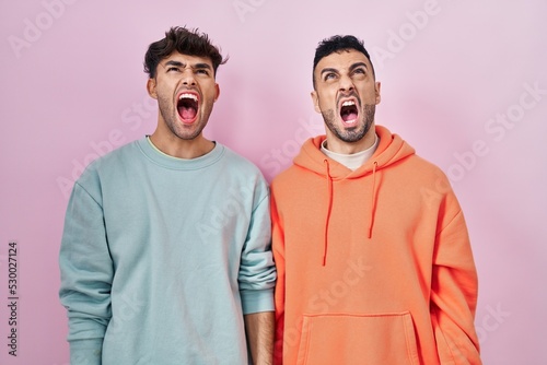 Young hispanic gay couple standing over pink background angry and mad screaming frustrated and furious, shouting with anger. rage and aggressive concept.