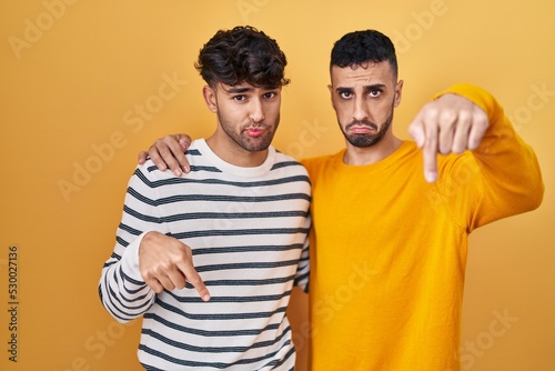 Young hispanic gay couple standing over yellow background pointing down looking sad and upset  indicating direction with fingers  unhappy and depressed.