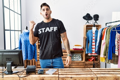 Young hispanic man working at retail boutique surprised pointing with hand finger to the side, open mouth amazed expression.