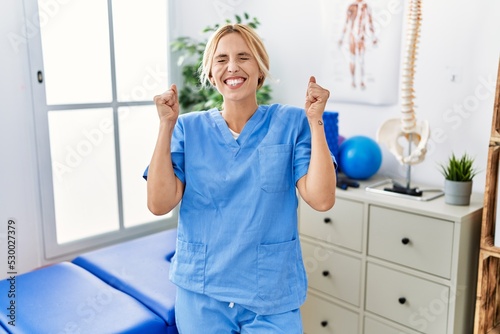 Beautiful blonde physiotherapist woman working at pain recovery clinic excited for success with arms raised and eyes closed celebrating victory smiling. winner concept.
