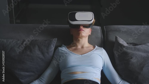 Woman in virtual reality goggles enters metaverse immersive experience via headset interface resting on sofa in minimalistic interior. Girl gaming in cyber space. Futuristic concept. photo