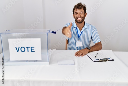 Young handsome man at political election sitting by ballot smiling friendly offering handshake as greeting and welcoming. successful business.