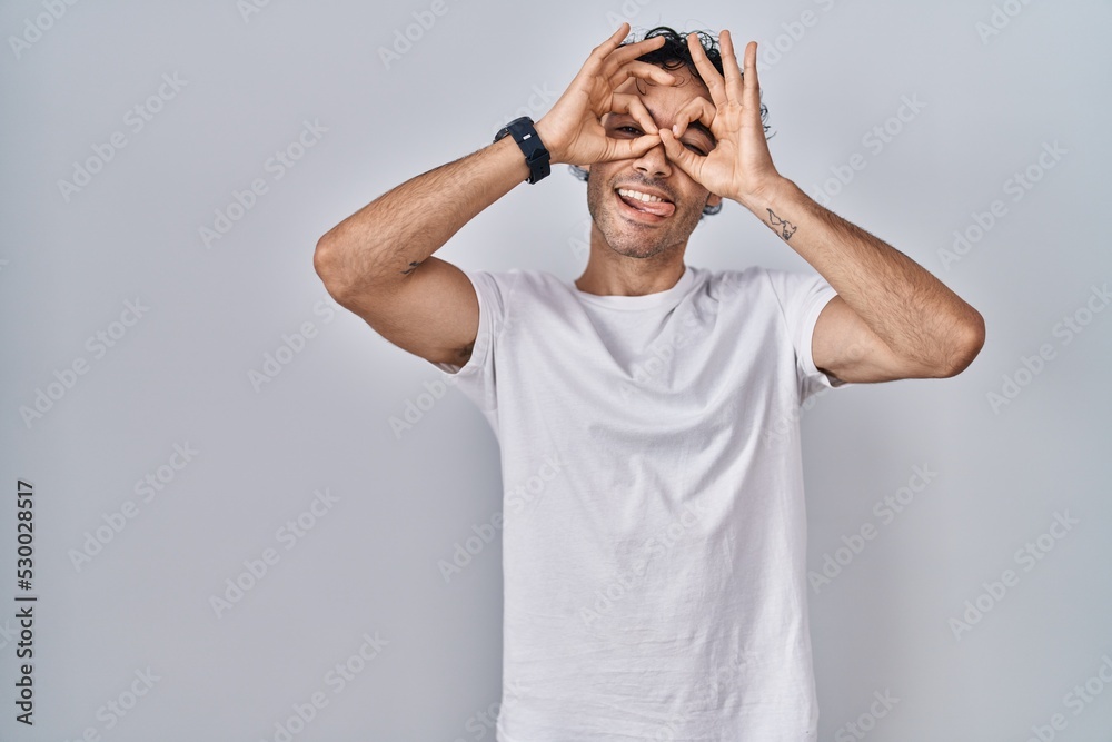 Hispanic man standing over isolated background doing ok gesture like binoculars sticking tongue out, eyes looking through fingers. crazy expression. Hispanic man standing over isolated background doing ok gesture like binoculars sticking tongue out, eyes looking through fingers. crazy expression.