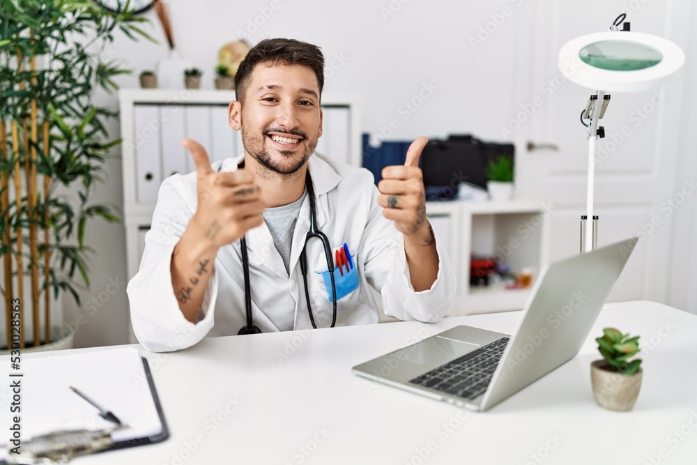 Young doctor working at the clinic using computer laptop success sign doing positive gesture with hand, thumbs up smiling and happy. cheerful expression and winner gesture.