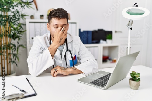 Young doctor working at the clinic using computer laptop tired rubbing nose and eyes feeling fatigue and headache. stress and frustration concept.