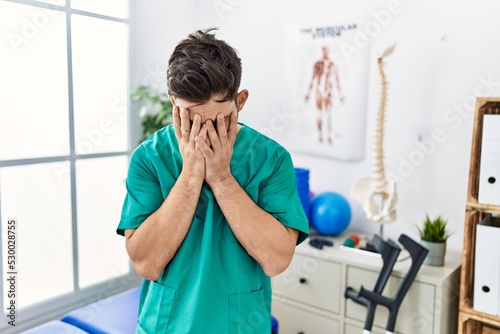 Young man with beard working at pain recovery clinic with sad expression covering face with hands while crying. depression concept.