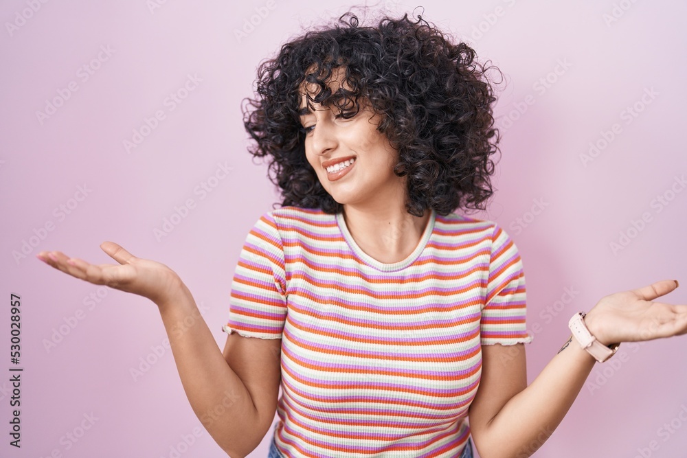 Young middle east woman standing over pink background smiling showing both hands open palms, presenting and advertising comparison and balance Young middle east woman standing over pink background smiling showing both hands open palms, presenting and advertising comparison and balance