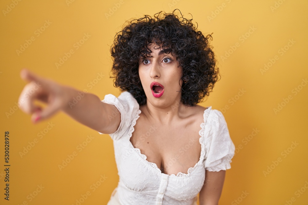 Young brunette woman with curly hair standing over yellow background pointing with finger surprised ahead, open mouth amazed expression, something on the front Young brunette woman with curly hair standing over yellow background pointing with finger surprised ahead, open mouth amazed expression, something on the front