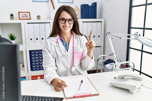 Young doctor woman wearing doctor uniform and stethoscope at the clinic showing and pointing up with finger number one while smiling confident and happy.