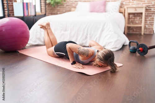 Young blonde woman doing yoga exercise on floor at bedroom