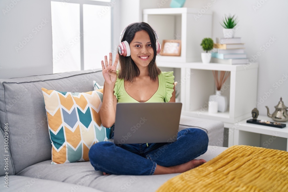 Hispanic young woman using laptop at home showing and pointing up with fingers number four while smiling confident and happy. Hispanic young woman using laptop at home showing and pointing up with fingers number four while smiling confident and happy.