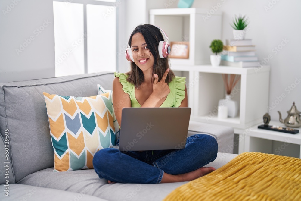 Naklejka premium Hispanic young woman using laptop at home cheerful with a smile of face pointing with hand and finger up to the side with happy and natural expression on face