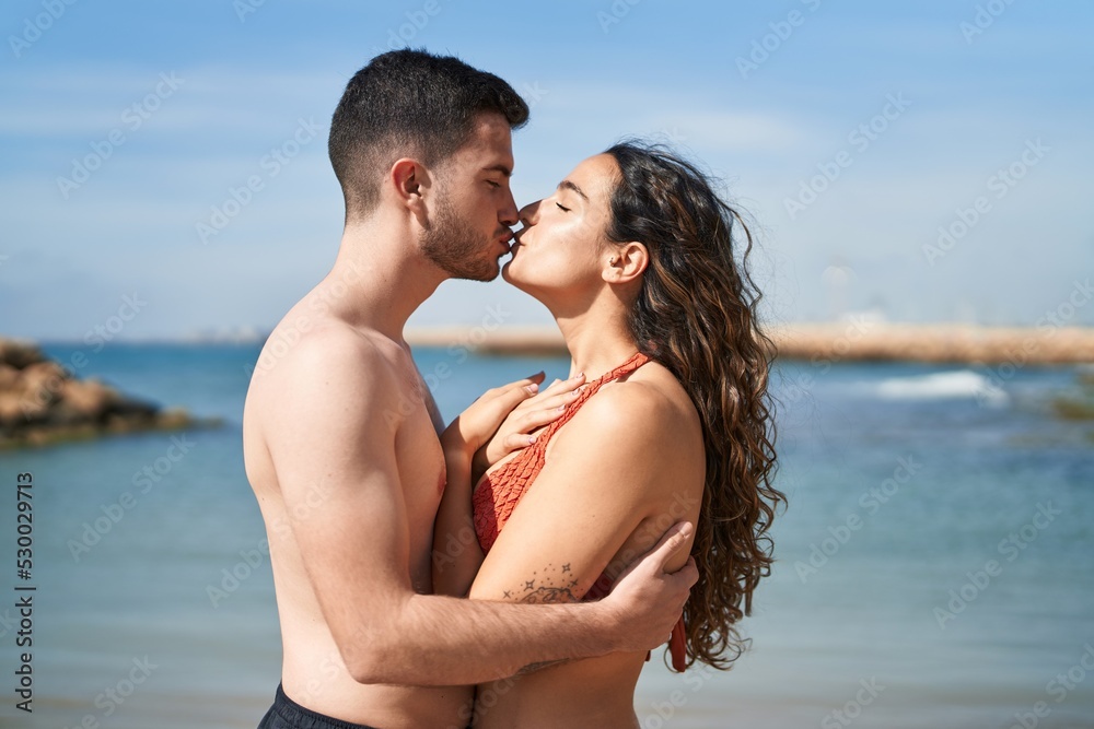 Young hispanic couple tourists wearing swimsuit hugging each other and kissing at seaside Young hispanic couple tourists wearing swimsuit hugging each other and kissing at seaside
