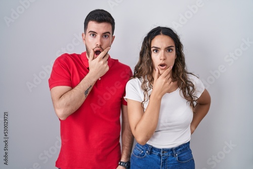 Young hispanic couple standing over isolated background looking fascinated with disbelief, surprise and amazed expression with hands on chin
