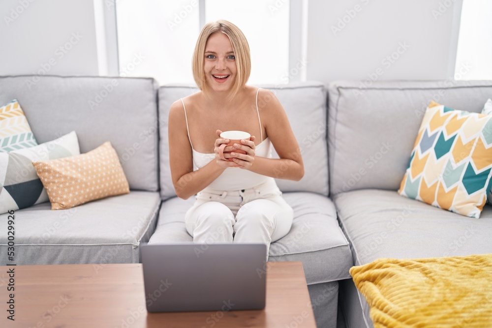 Young blonde woman using laptop and drinking coffee sitting on sofa at home Young blonde woman using laptop and drinking coffee sitting on sofa at home