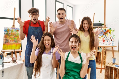 Group of five hispanic artists at art studio celebrating mad and crazy for success with arms raised and closed eyes screaming excited. winner concept