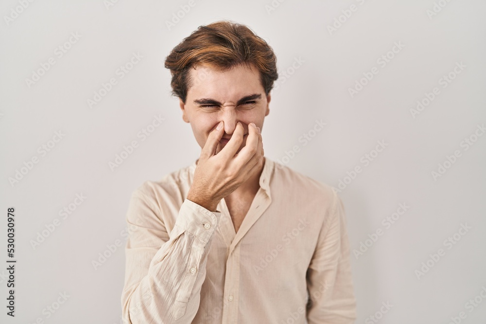 Young man standing over isolated background smelling something stinky and disgusting, intolerable smell, holding breath with fingers on nose. bad smell Young man standing over isolated background smelling something stinky and disgusting, intolerable smell, holding breath with fingers on nose. bad smell