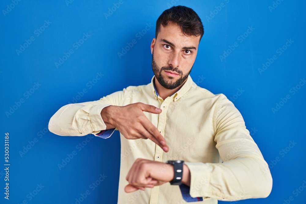 Handsome hispanic man standing over blue background in hurry pointing to watch time, impatience, upset and angry for deadline delay