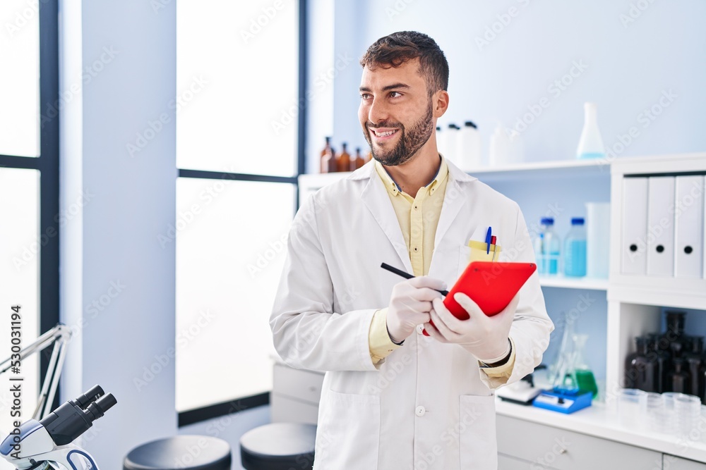 Young hispanic man wearing scientist uniform using touchpad at laboratory