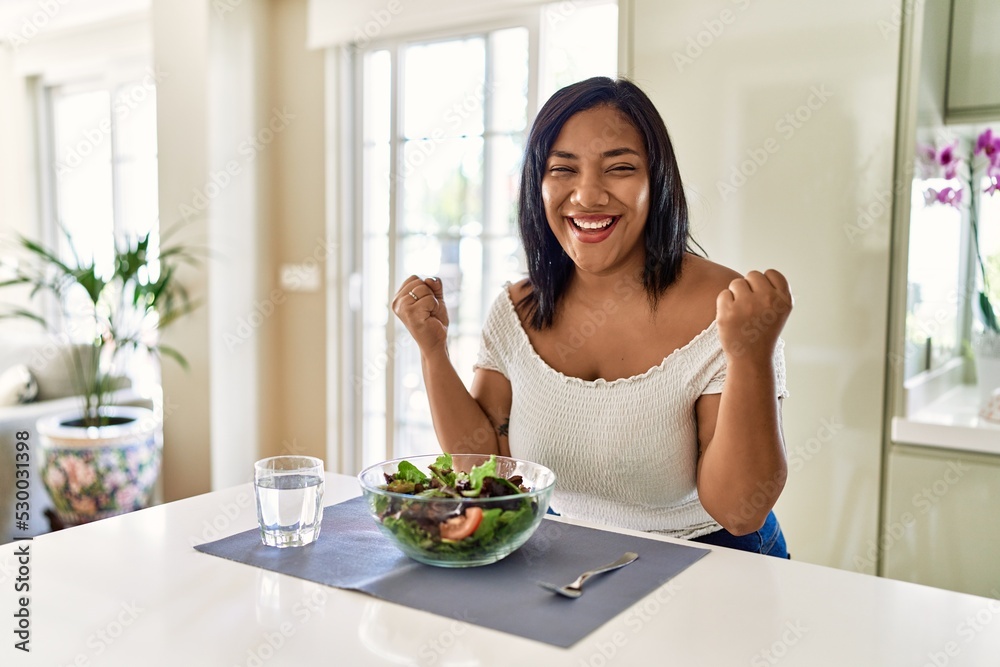 Young hispanic woman eating healthy salad at home celebrating surprised and amazed for success with arms raised and open eyes. winner concept. Young hispanic woman eating healthy salad at home celebrating surprised and amazed for success with arms raised and open eyes. winner concept.