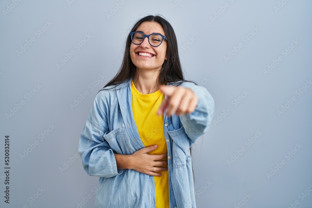 Young hispanic woman standing over blue background laughing at you, pointing finger to the camera with hand over body, shame expression Young hispanic woman standing over blue background laughing at you, pointing finger to the camera with hand over body, shame expression