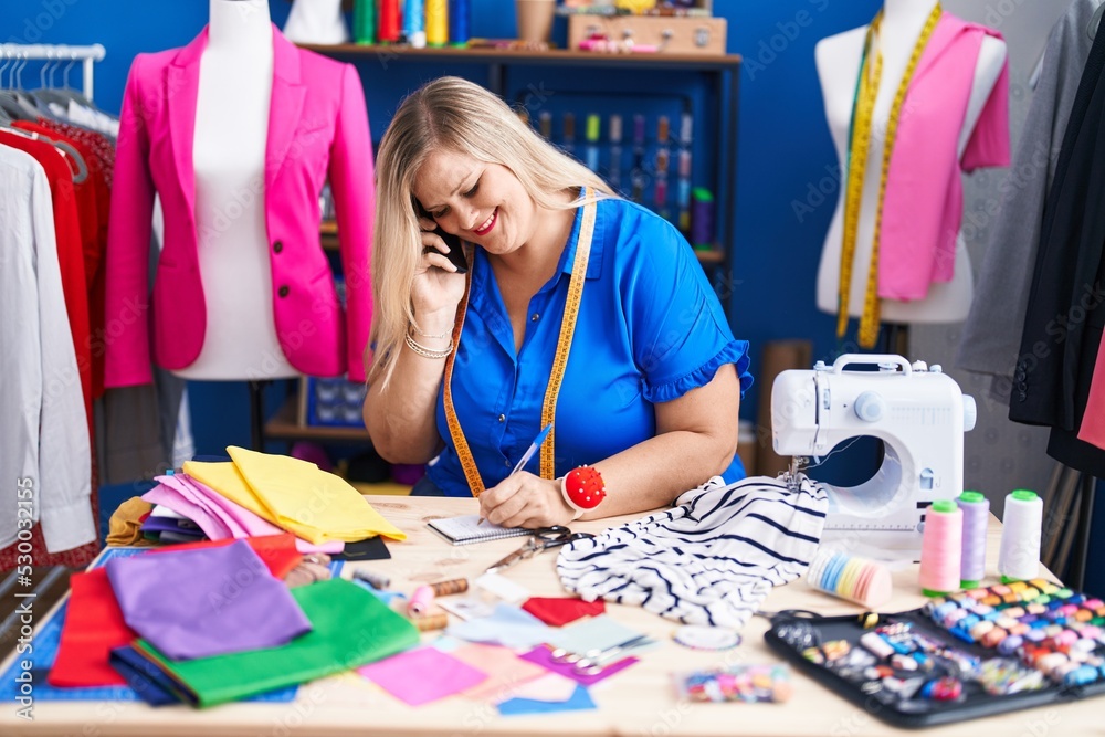 Fototapeta premium Young woman tailor talking on smartphone writing on notebook at sewing studio