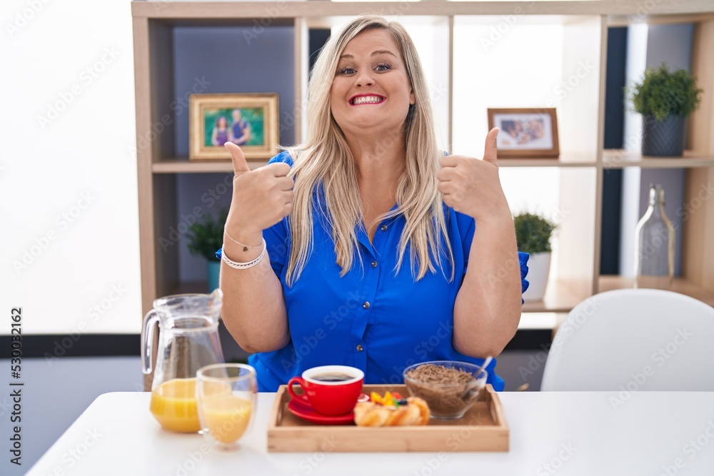 Caucasian plus size woman eating breakfast at home success sign doing positive gesture with hand, thumbs up smiling and happy. cheerful expression and winner gesture.