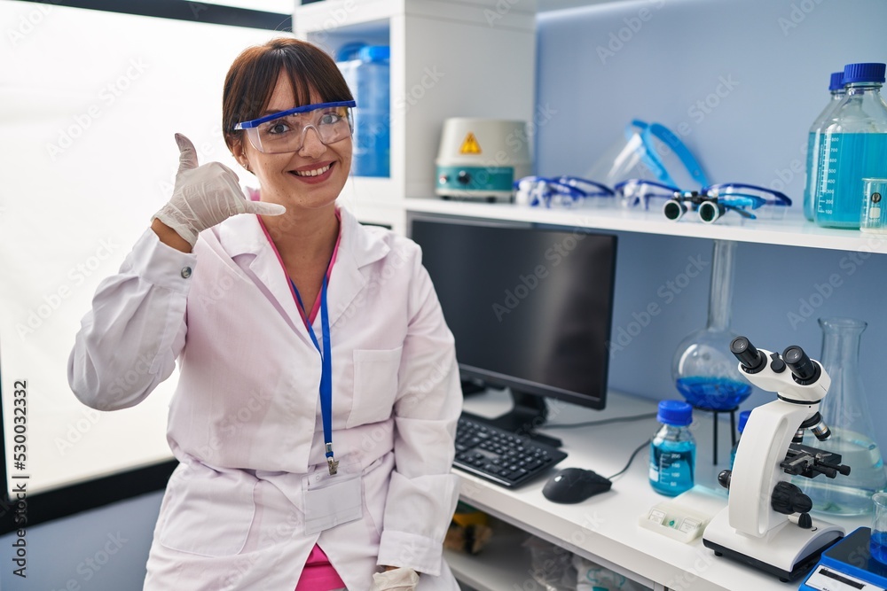 Young brunette woman working at scientist laboratory smiling doing phone gesture with hand and fingers like talking on the telephone. communicating concepts. Young brunette woman working at scientist laboratory smiling doing phone gesture with hand and fingers like talking on the telephone. communicating concepts.
