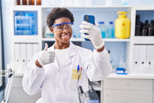 African american woman working at scientist laboratory doing video call with smartphone smiling happy and positive, thumb up doing excellent and approval sign