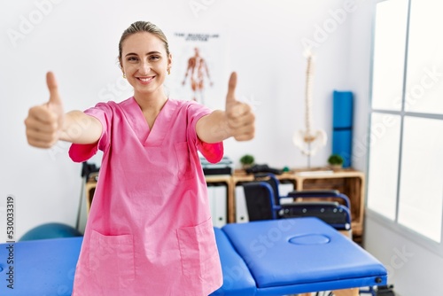 Young blonde woman working at pain recovery clinic approving doing positive gesture with hand  thumbs up smiling and happy for success. winner gesture.
